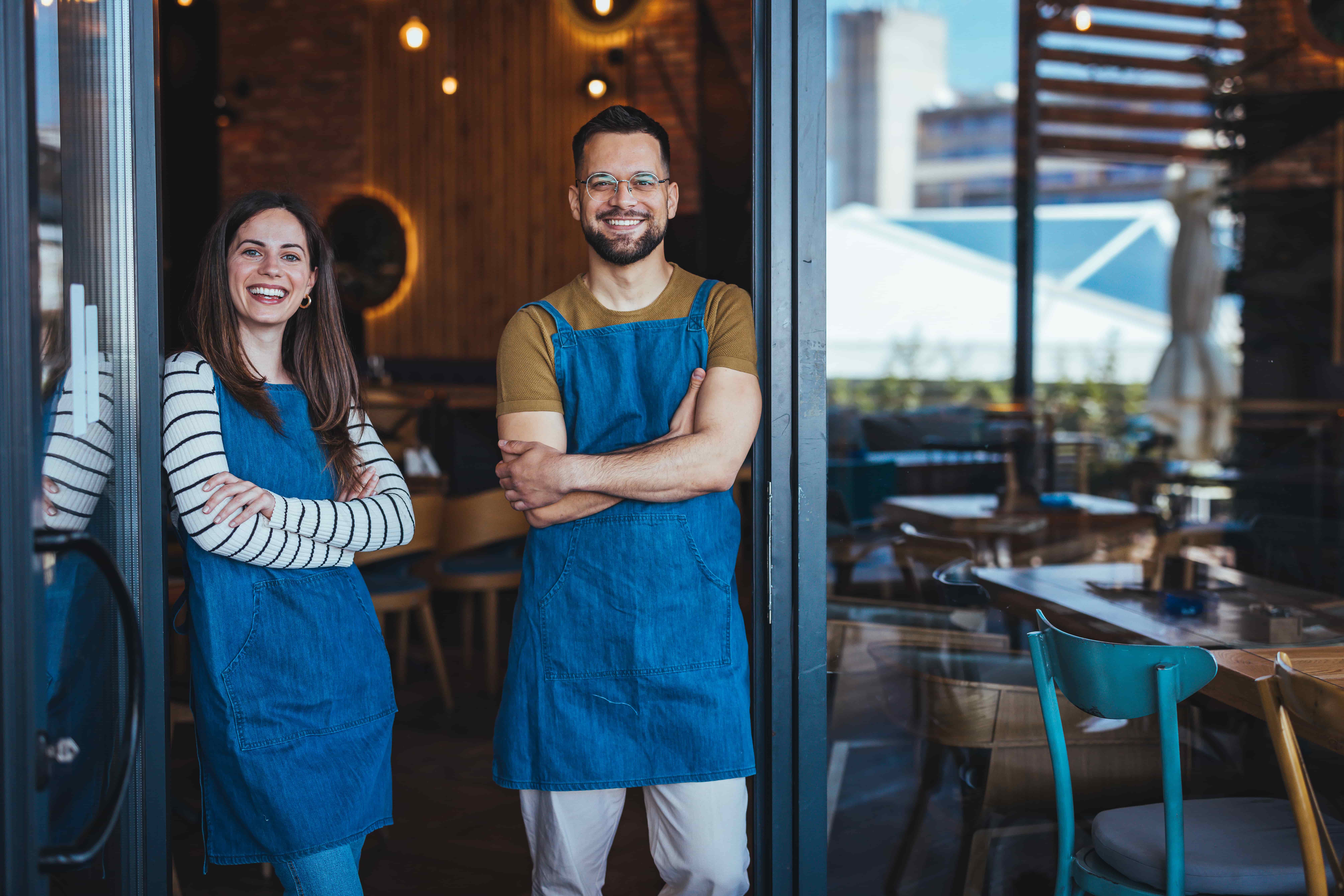 Smiling Café Owners Standing Proudly at Their Restaurant Entrance_1063402776-min